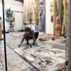 Iva Geuorguieva, a young woman, is pictured in an art studio. She is kneeling down close to the floor where she's drawing on a large black and white art piece. She wears a happy expression.