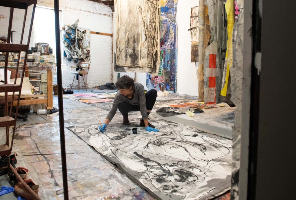Iva Geuorguieva, a young woman, is pictured in an art studio. She is kneeling down close to the floor where she's drawing on a large black and white art piece. She wears a happy expression.