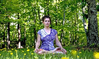 A young woman meditating in the middle of a meadow of yellow flowers surrounded by a lush forest. She is sitting cross legged with her arms and hands are relaxed at her sides, and she has a calm expression with her eyes closed.